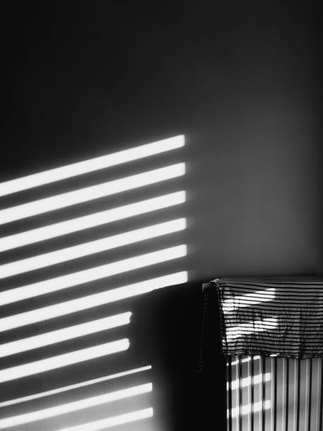 istock Sunlight is streaming through blinds, creating stripes of light and shadow on wall and radiator with striped cloth draped over it, in black and white composition. Lines of light and shadow on wall 2203659543