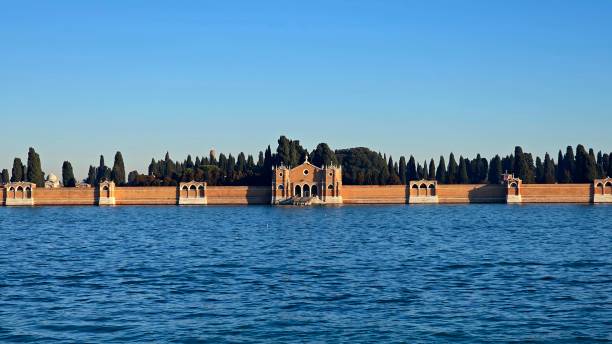 Close view of San Michele, Venice's cemetery island. The walled complex features arched gateways, a central entrance with twin towers, and tall cypress trees, all surrounded by calm waters.