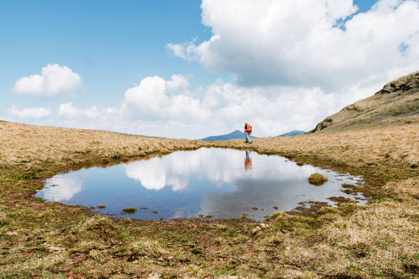 Hiker crossing a small body of water in an open, grassy landscape. The person is wearing outdoor clothing and carrying a backpack. Their reflection is visible in the calm water, creating a beautiful mirror effect. The sky is bright blue with scattered white clouds, and the surroundings look peaceful and natural.