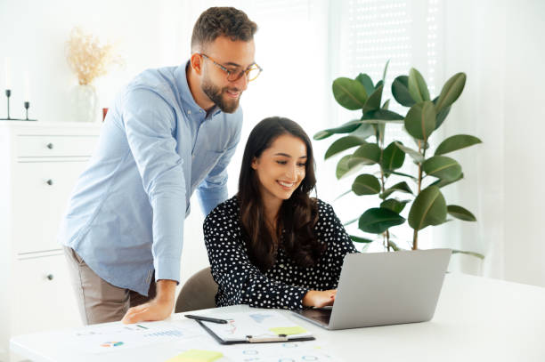 istock Man assisting female colleague while working on a laptop at a desk, professional teamwork in a modern office, positive coworkers working together on a project, developing ideas and business plan 2203702385