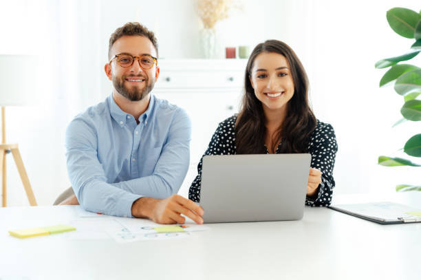 istock Positive multiracial business colleagues sitting side by side at a work desk in a modern office space with a laptop in front, working together on a new project, looking at the camera, smile friendly 2203705742
