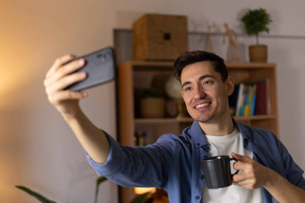 istock Smiling young man taking a selfie with his smartphone while holding a coffee mug in his living room 2203721989