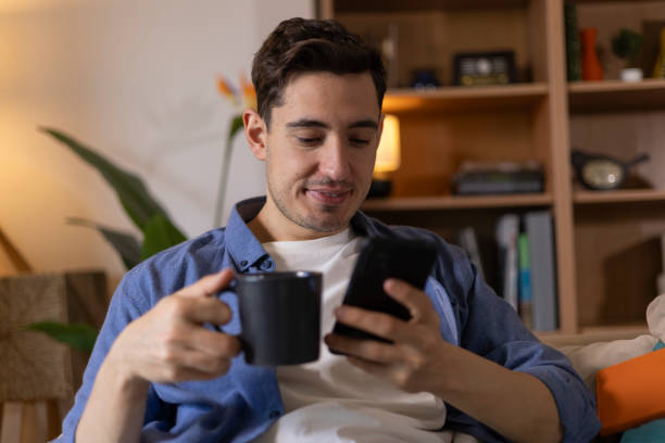istock Smiling young man enjoying a cup of coffee while using his smartphone on the sofa at home 2203722077