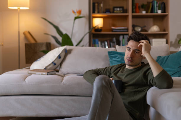 istock Young man sitting on the floor by his sofa, holding a mug and lost in thought 2203722149