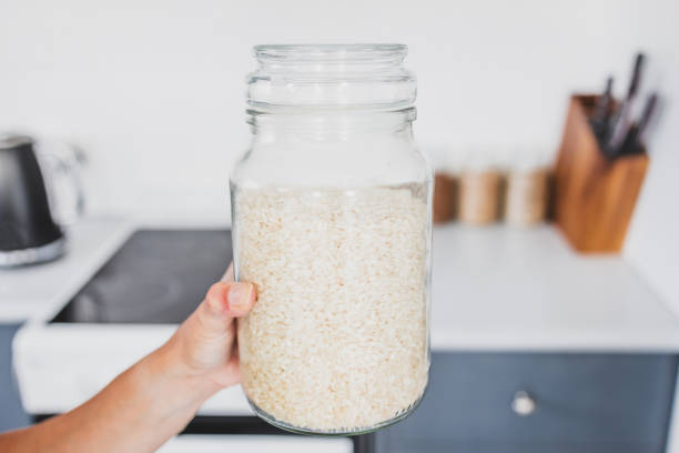 sosteniendo de mano un frasco de vidrio de arroz arborio frente al bokeh de la cocina, cocinando desde cero y teniendo una despensa organizada - arroz arborio fotografías e imágenes de stock