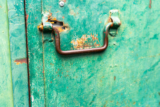 A close-up of an old, rusted metal handle attached to a weathered green door. The peeling paint, corrosion, and rough textures emphasize the passage of time and exposure to the elements. The mix of green, brown, and orange tones creates a visually striking contrast, while the scratches and dents add character to the industrial aesthetic.