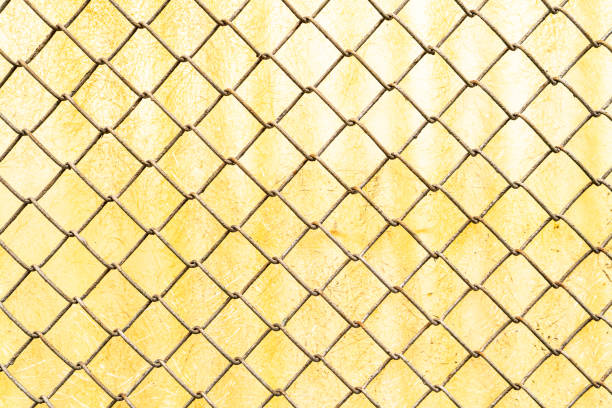 A close-up of a chain-link fence set against a weathered yellow surface, creating a striking geometric pattern. The metal wires show signs of rust and age, adding texture and depth to the composition. The contrast between the rigid structure of the fence and the scratched, worn background creates an industrial yet artistic aesthetic.