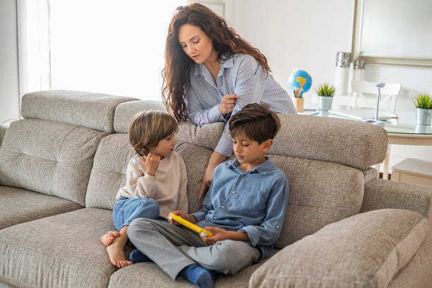 istock Mother is watching her two young sons playing a video game together on a comfortable sofa in their living room, creating a heartwarming scene of family bonding 2203739211