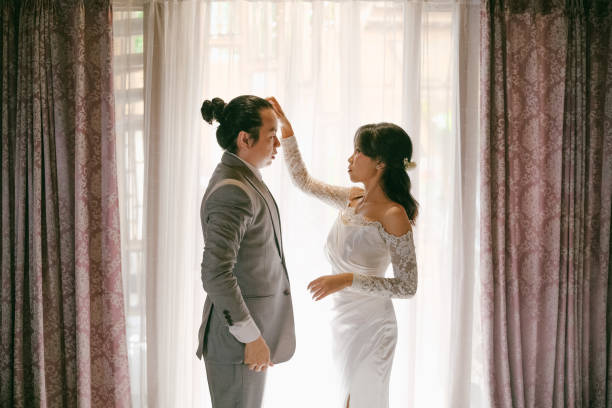 A heartfelt moment between a bride and groom as she gently fixes his hair, standing in front of softly lit curtains.