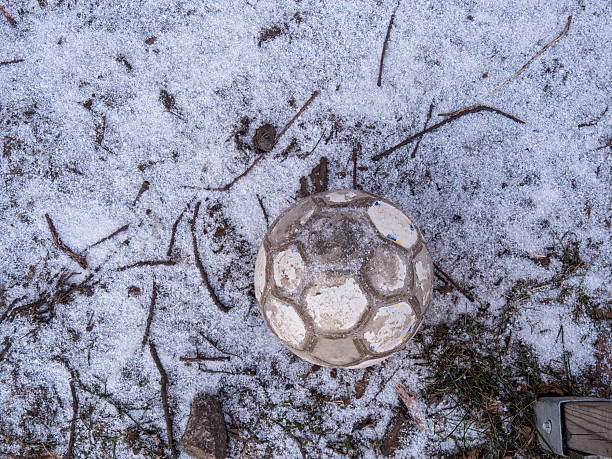 A worn soccer ball on a snowy ground with scattered twigs and grass.