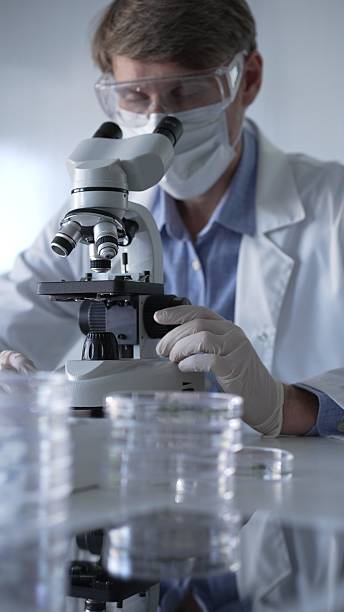 istock Man scientist wearing lab coat, white gloves, medical mask and protective glasses using microscope doing research in modern laboratory, portrait view. Low key lighting photo. Science and Medicine 2203759113