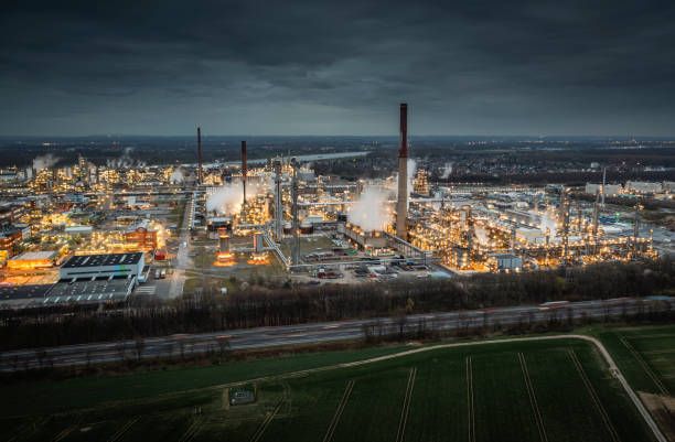 Aerial view of a sprawling industrial refinery at dusk, with bright lights illuminating the complex network of factories, smokestacks, and pipelines. Highway in the foreground.