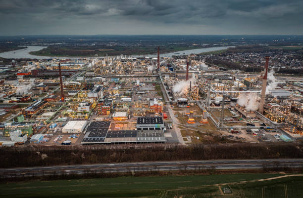 Aerial view of a massive industrial refinery complex at dusk, featuring multiple smokestacks emitting steam, interconnected pipelines, and large manufacturing facilities. The intricate infrastructure and bright lights showcase the complexity of modern energy production and heavy industry.