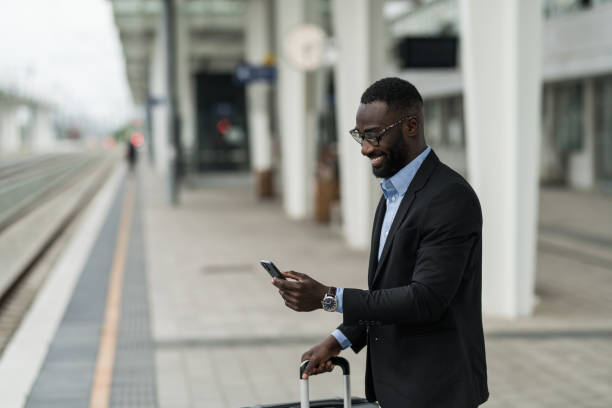 istock Cheerful black businessman using smart phone while waiting for train on railway station. 2203765375