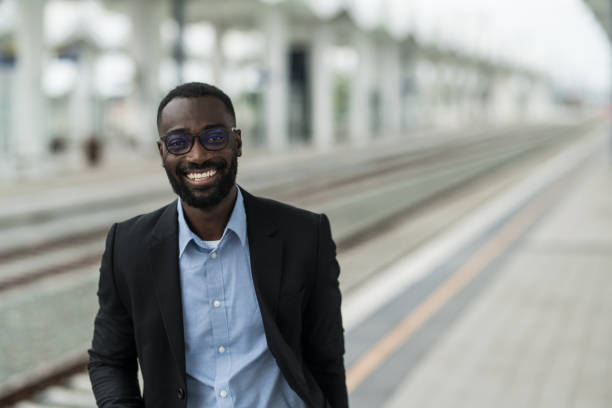 istock Cheerful black businessman looking at camera while waiting for train on railway station. 2203765831