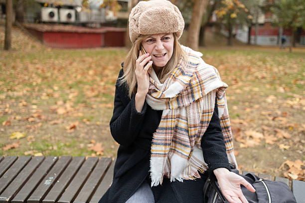 Angry woman wearing winter clothes complaining on the phone while sitting on a bench in a park during autumn