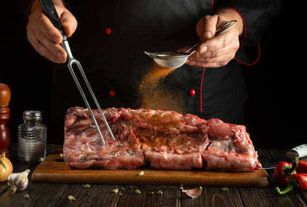 istock A chef meticulously seasons a large cut of meat using a strainer and a fork. Fresh herbs and spices are prominently present on the wooden board 2203782793