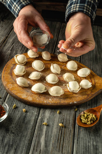 istock Hands skillfully sprinkle salt over freshly shaped dumplings arranged on a wooden board. The warm kitchen atmosphere is enhanced by wooden textures and scattered ingredients, creating a homely feel 2203783082