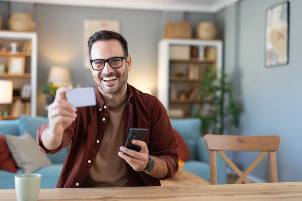 istock Smiling man holds a card while using his smartphone at home. 2203784988