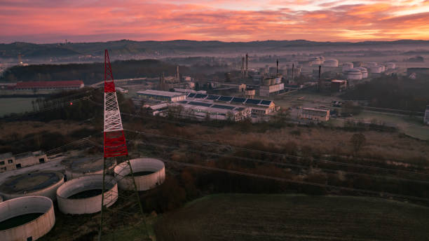 Drone view of industrial complex and power lines under a vibrant sunset sky. The scene includes factories, storage tanks, and a colorful sky transitioning from orange to purple.