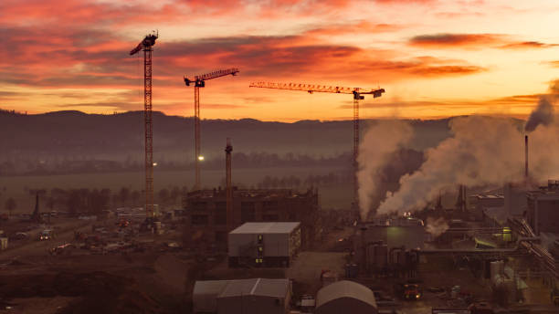 Aerial view of an industrial site with towering cranes and smoke rising against a vibrant sunset sky. The landscape features silhouetted hills and open fields, capturing the essence of industry at dusk.