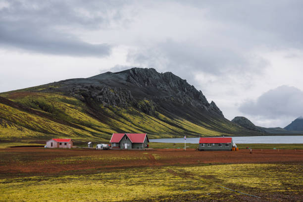 A peaceful Icelandic highland settlement nestled at the foot of rugged mountains, overlooking a tranquil lake. The scattered houses with red roofs contrast against the lush green slopes and volcanic terrain. A flag waves gently in the breeze, emphasizing the isolation and serenity of this remote wilderness