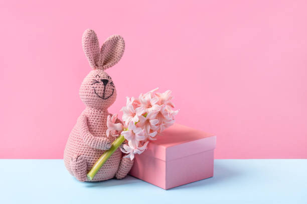 Gift box, knitted toy pink bunny with Easter egg and bouquet of spring flowers on blue table and purple background. Concept of spring, Easter, Mother's Day