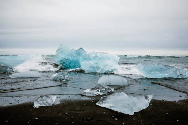 A mesmerizing view of Diamond Beach in Iceland, where crystal-clear icebergs from the Jökulsárlón Glacier Lagoon wash ashore on the striking black sand. The jagged ice formations glisten under the overcast sky as waves crash onto the volcanic shoreline, creating a dramatic contrast between the icy blue fragments and the dark sand. This breathtaking landscape is a must-visit destination for nature lovers, photographers, and travelers seeking the raw beauty of Iceland’s ever-changing environment.