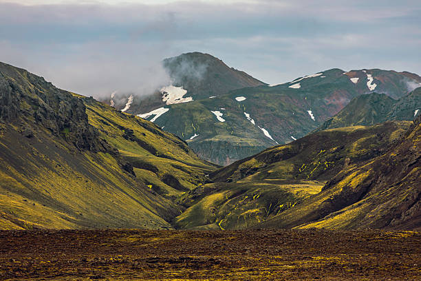 A breathtaking view of the Icelandic Highlands, where moss-covered volcanic hills lead up to towering mountains partially shrouded in mist. Patches of snow cling to the rugged slopes, adding contrast to the lush green valley. The dramatic landscape captures the raw beauty and untouched wilderness of Iceland’s remote interior