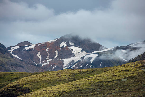 A stunning view of Iceland’s remote highlands, where rugged mountains with patches of snow and glacial ice rise above lush green valleys. Mist drifts over the slopes, creating a mystical atmosphere that enhances the dramatic beauty of this untouched wilderness. The contrast between the dark volcanic rock, white snow, and green moss-covered hills highlights the country’s diverse landscapes. This breathtaking scenery is a haven for adventurers and photographers seeking Iceland’s raw, natural beauty.