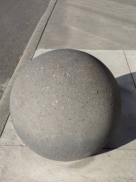 A large concrete sphere positioned on a paved urban sidewalk. The setting features clean lines and a bright daylight ambiance, depicting a minimalistic urban design element.