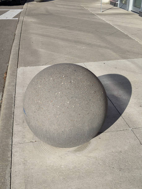 A large concrete sphere positioned on a paved urban sidewalk. The setting features clean lines and a bright daylight ambiance, depicting a minimalistic urban design element.