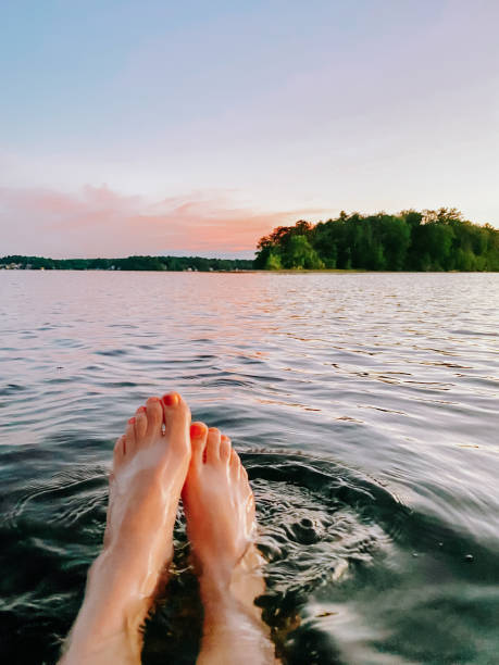 A serene moment of wild swimming, captured with feet floating in a tranquil lake at sunset. The golden hues reflect on the rippling water, while the lush tree-lined shore in the distance adds to the peaceful atmosphere