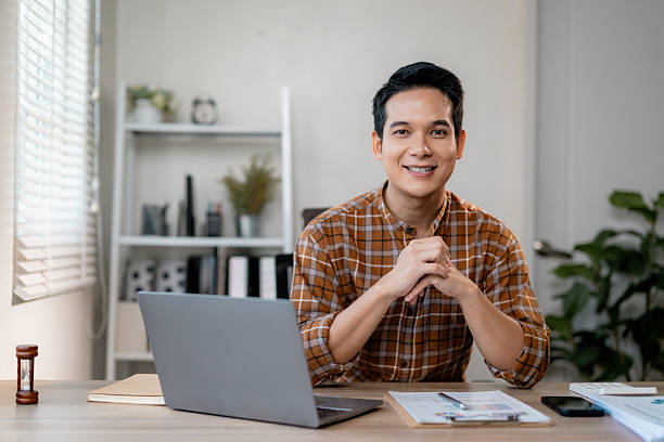 istock Young Asian freelancer working remotely, smiling confidently near laptop, home office setting with scattered documents, green potted plants nearby 2203822046