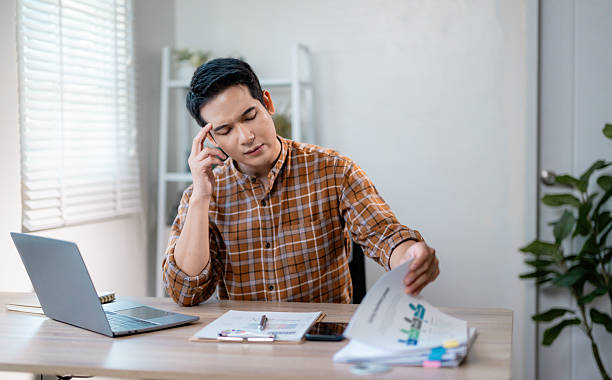 istock Overwhelmed asian professional experiencing work-related stress, clutching head while multitasking on phone and laptop in home workspace, displaying mental exhaustion 2203822063