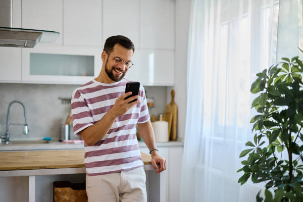 istock Young man using smartphone and smiling in kitchen at home 2203840331