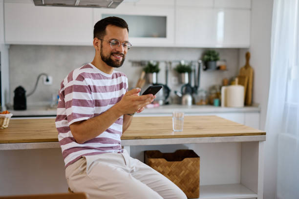 istock Young man using smartphone and smiling in kitchen at home 2203840332