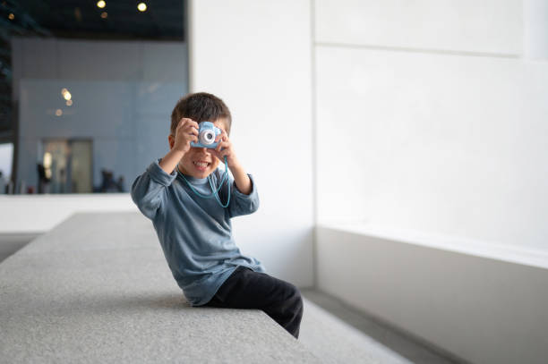 A young boy joyfully poses with his camera, capturing his surroundings while sitting on a modern ledge in a minimalist environment with soft natural light streaming in.