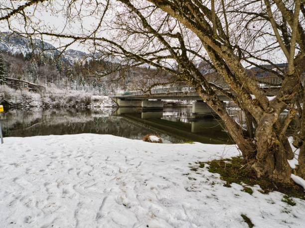 Snow-covered landscape with a bridge over a calm river, surrounded by bare trees and mountains in the background.