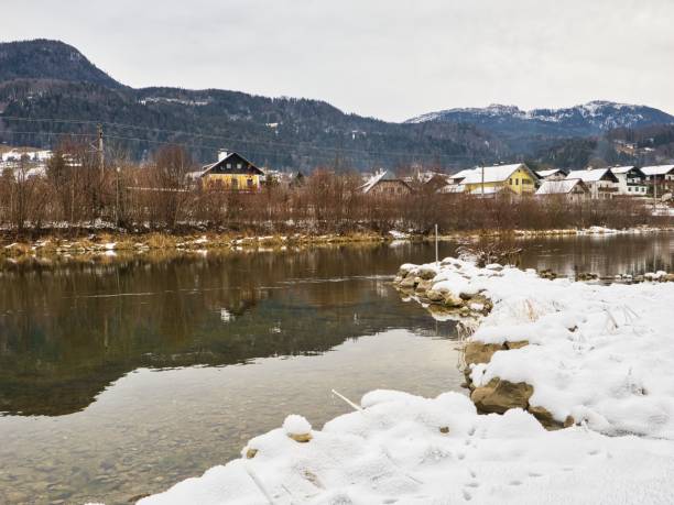 istock Winter village by a calm river with mountain backdrop. 2203855844