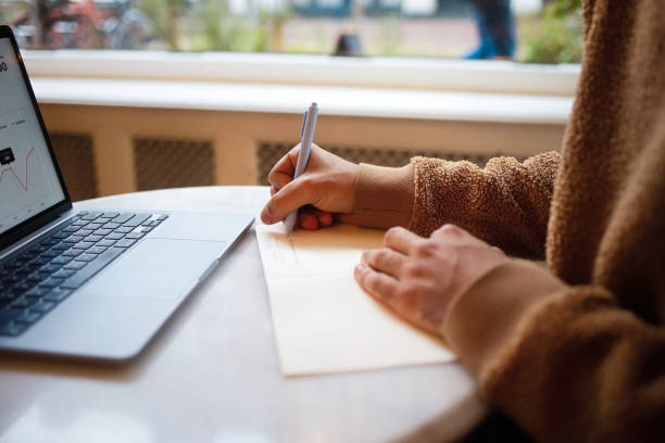istock Person Writing Notes in a Notebook Beside Laptop in Cafe 2203869469