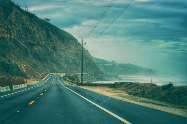A scenic stretch of Highway 1 on the West Coast of California, where the road winds between rugged cliffs and the Pacific Ocean. The misty atmosphere and dramatic clouds enhance the breathtaking coastal views, making this an iconic drive for travelers exploring California’s stunning shoreline.