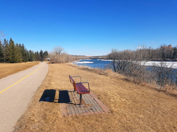 Park Bench looking to Bow River. Baker Park NW. Calgary.  Group of people on path in background. Ice and Snow in River.