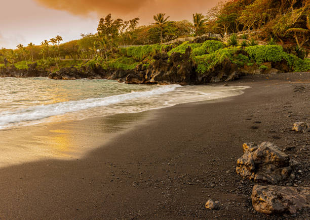Black sand beach at Waianapanapa State Park, Maui, Hawaii, with tropical vegetation, volcanic rocks and turquoise Pacific Ocean waves.