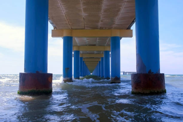 Sea under pier among parallel blue columns making narrow corridor in water. Seascape through pier with sea waves and blue sky at sunny day.