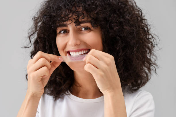 istock Young woman applying whitening strip on her teeth against light grey background 2203909797