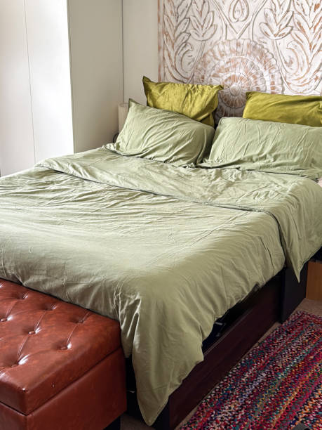 istock Image of double bed with co-ordinated green bedding, pillow and cushion stack, folded duvet, button backed, leather ottoman focus on foreground, soft home furnishing 2203919548