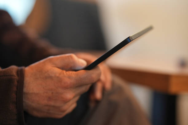 A close-up of a person holding a tablet in one hand while seated in a comfortable indoor setting. The blurred background gives a soft, warm ambiance.