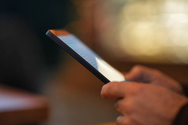 A close-up of a person holding a tablet with the screen displaying text. The background is softly blurred with warm lighting, creating a cozy reading atmosphere.