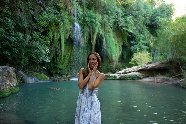 A joyful woman in a white dress stands by a scenic river with a waterfall in the background, expressing excitement and happiness in a lush natural setting.